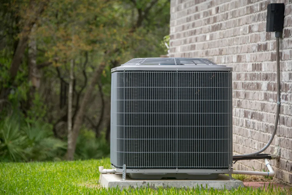 Outdoor AC unit positioned beside a brick wall, surrounded by grass and trees, illustrating proper clearance for air intake and output.