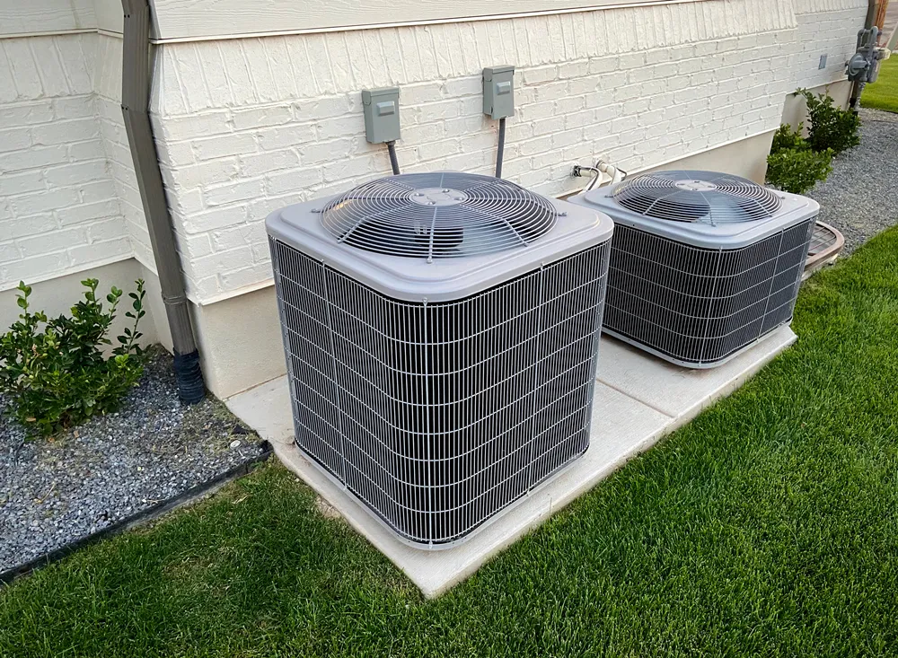 Outdoor AC unit clearance with two air conditioning units positioned on a concrete pad beside a residential building, surrounded by grass and shrubs, illustrating proper HVAC system maintenance.