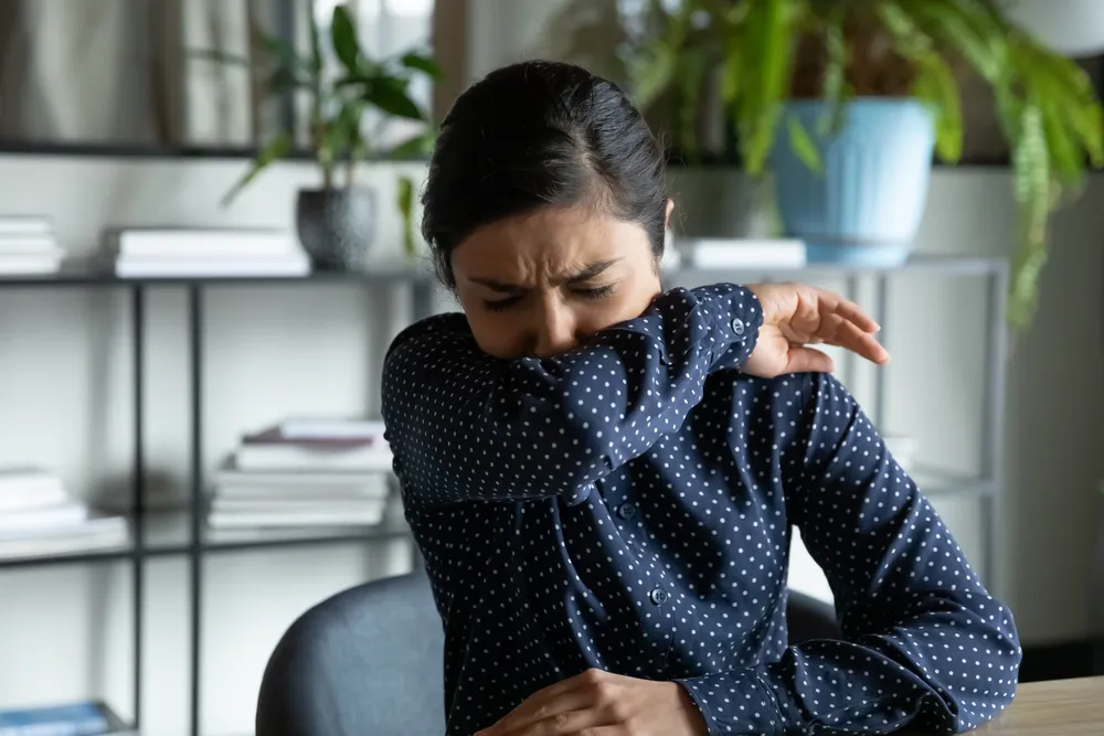 Woman sneezing into her elbow while sitting at a desk, illustrating allergy symptoms in an indoor setting with plants and bookshelves.