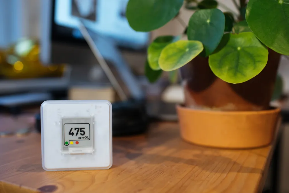 Indoor air quality monitor displaying 475 ppm CO₂, with a potted plant in the background, illustrating winter air quality concerns.