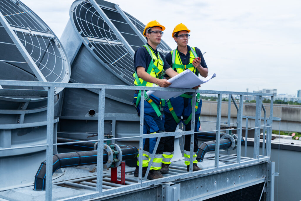 Engineers reviewing HVAC blueprints on a rooftop, wearing safety gear and hard hats, overseeing air conditioning units in Scottsdale, AZ.