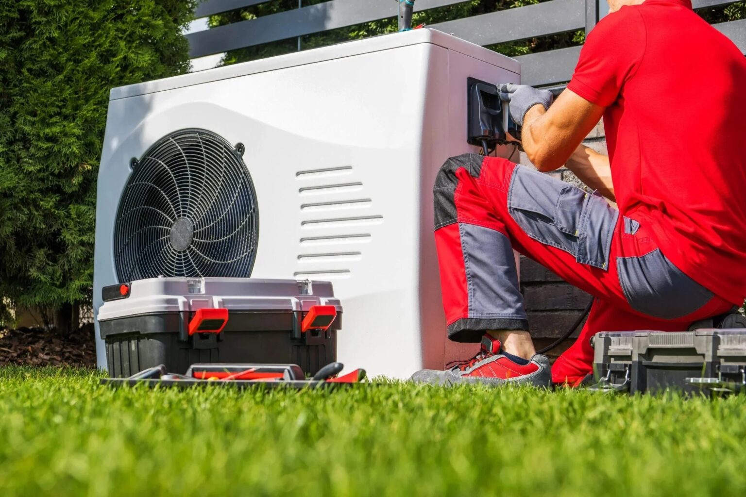 A technician kneeling at an outdoor heat pump on a lawn, his toolbox open beside him.