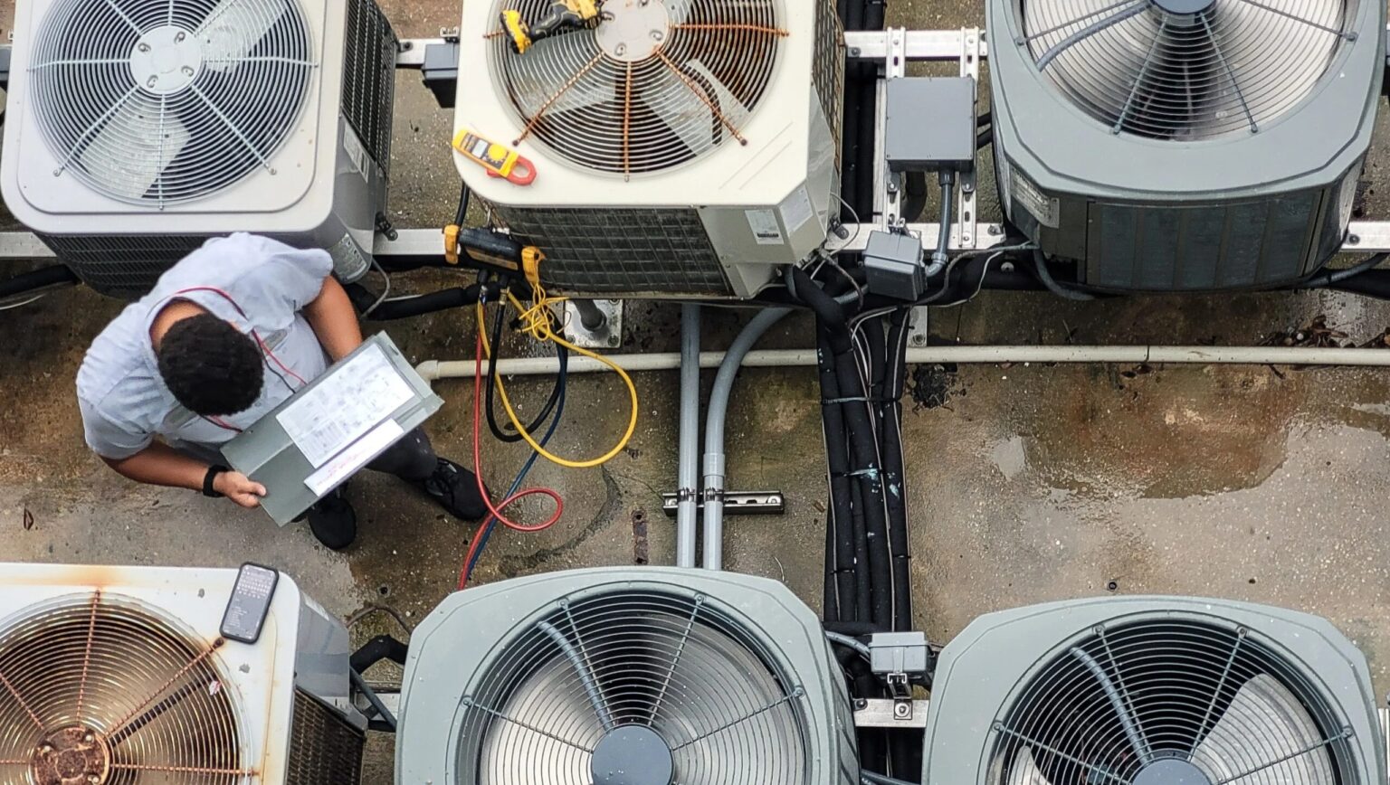An HVAC technician working on a laptop among rows of outdoor units, seen from above.