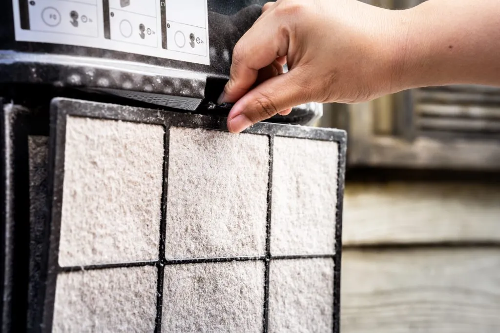 A hand lifting a dirty pleated filter out of an air purification unit.