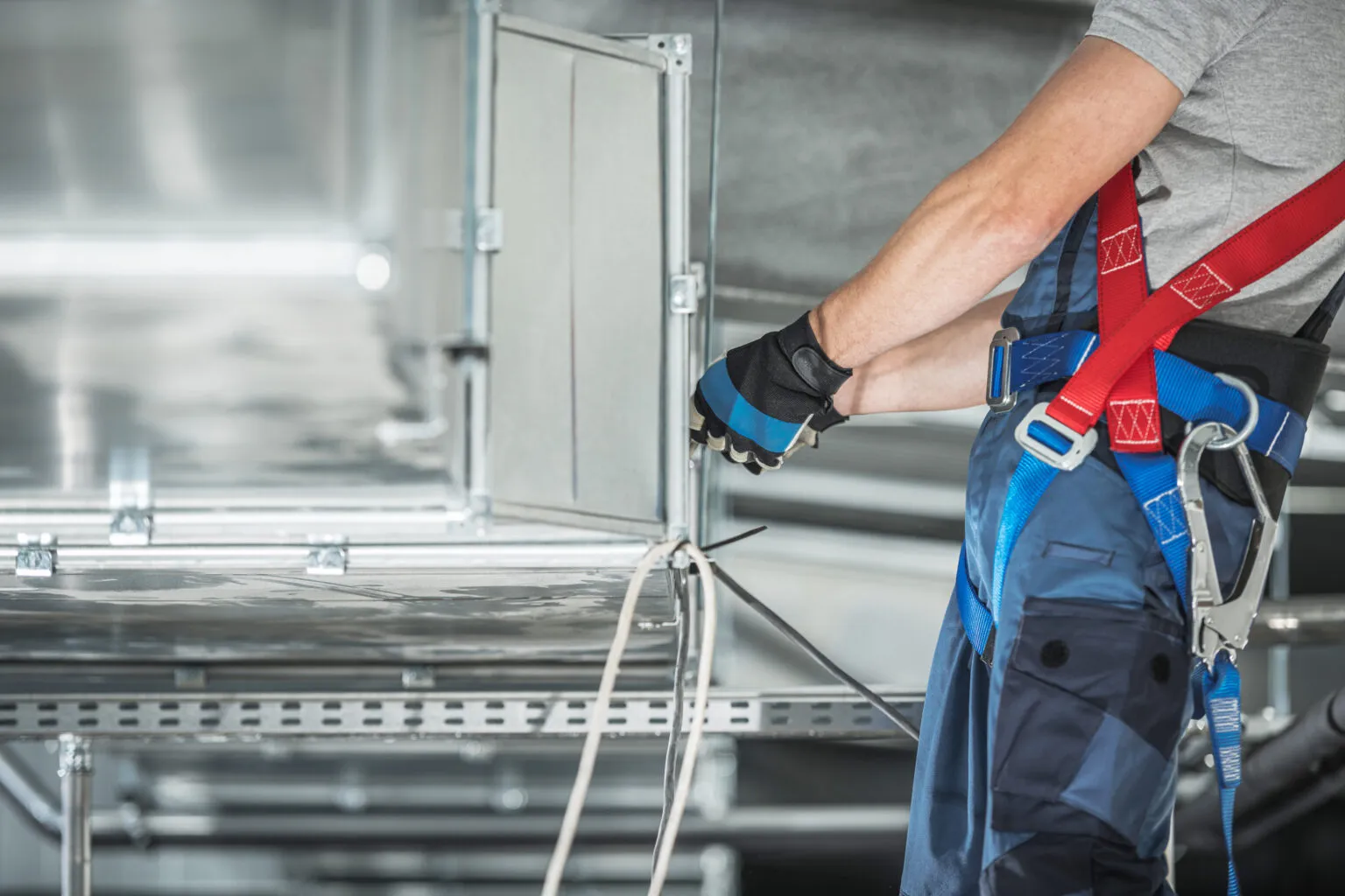 A technician in a safety harness working on ventilation ductwork in a plant room.