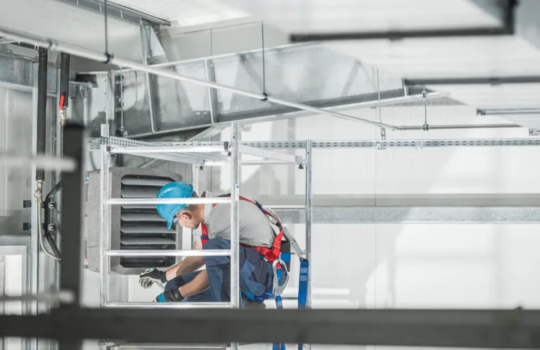 A commercial HVAC technician working at height on ductwork inside a warehouse.
