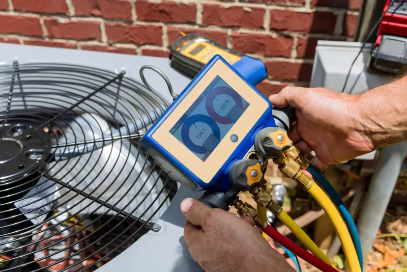 Hands using a digital HVAC gauge on an air conditioning unit, measuring pressure levels, with a fan and brick wall in the background, relevant to cooling efficiency tips for Scottsdale homeowners.
