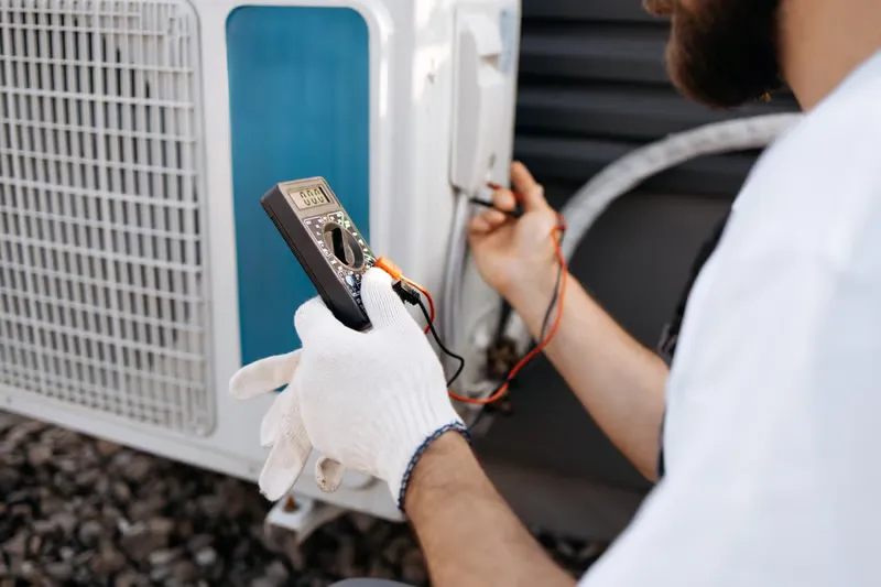 Technician testing air conditioning unit with multimeter, wearing gloves, assessing HVAC performance issues in Mesa, AZ.