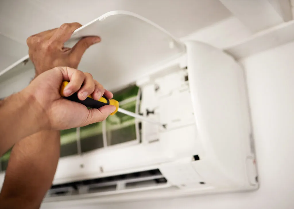 A technician's hands servicing the open panel of an air conditioning unit.