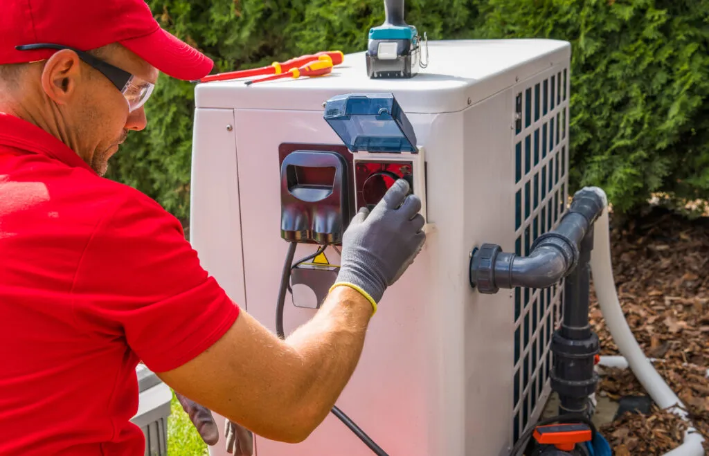A heating technician servicing an outdoor heat pump unit beside a garden.